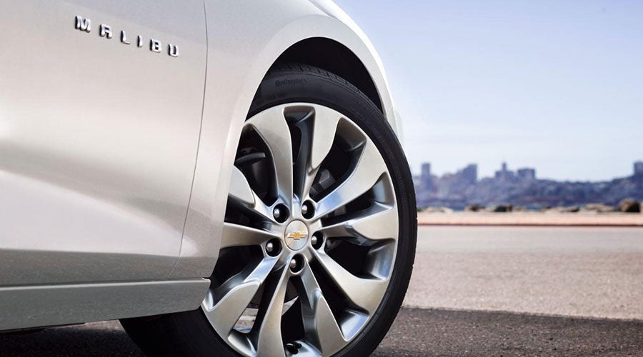 Side view of a white Chevrolet Malibu car, showing its chrome wheel, tire, and "MALIBU" emblem, with a city skyline in the background.