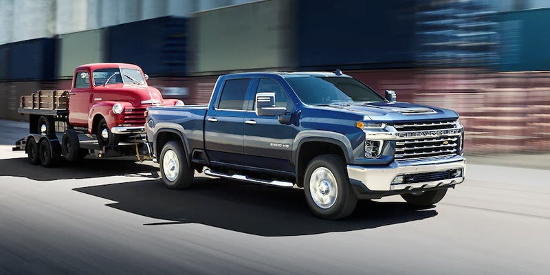 Blue Chevy Silverado towing a vintage red pickup on a trailer, moving quickly on a road.
