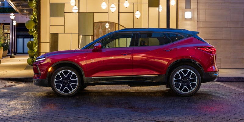Side view of a metallic red SUV with a black roof, parked on a street with a modern building and lights in the background.