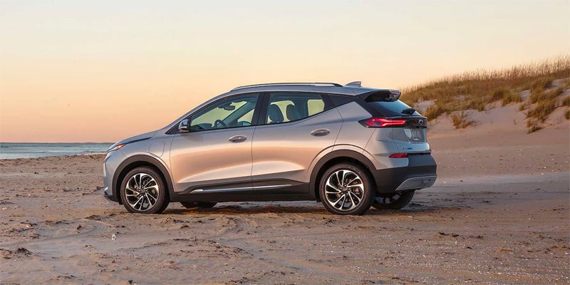 Silver SUV parked on a sandy beach with the ocean and dunes in the background at sunset.