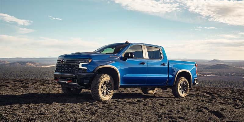 Blue Chevrolet Silverado ZR2 pickup truck parked on a rocky hilltop overlooking a vast forested landscape under a cloudy sky.