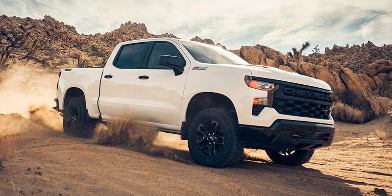 White Chevrolet pickup truck driving off-road on a dusty trail in a rocky desert landscape.