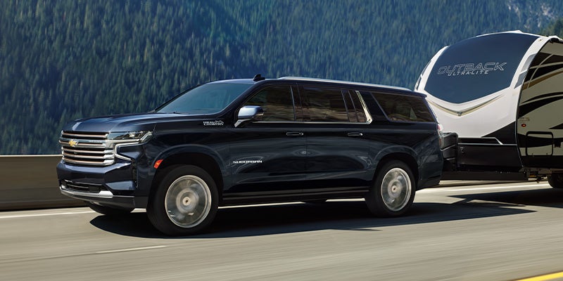 Dark blue Chevy Suburban SUV towing a white travel trailer on a highway with a forested mountain in the background.