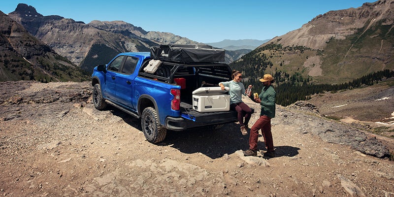 Blue pickup truck with rooftop tent parked on a rocky mountain pass. Two people are by the tailgate with a cooler.
