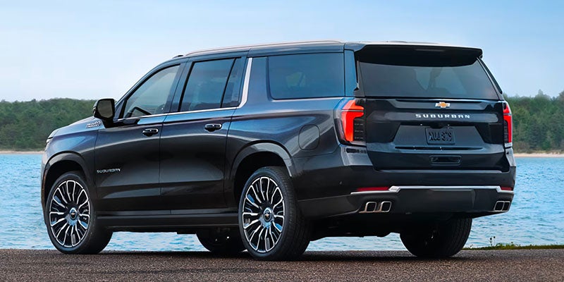 Black Chevrolet Suburban SUV, rear passenger side view, parked by water with trees in the background.