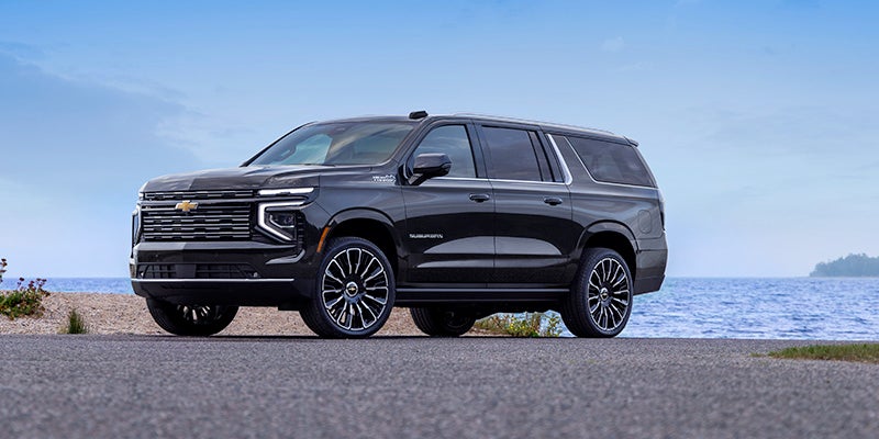 Dark grey Chevrolet Suburban SUV parked on asphalt by a calm blue lake under a clear sky.