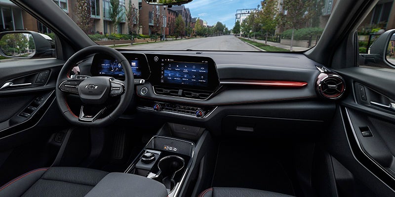 Car interior showing the dashboard, steering wheel, and dual digital displays with a road and buildings visible outside.