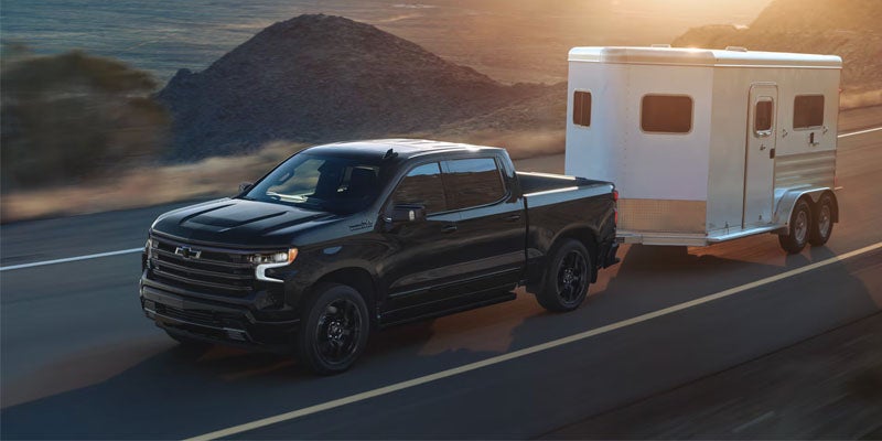 Black pickup truck towing a white horse trailer on a highway with mountains and sunset in the background.
