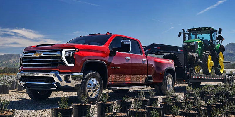 Red Chevrolet 3500 HD pickup truck towing a green tractor on a flatbed trailer through a nursery.