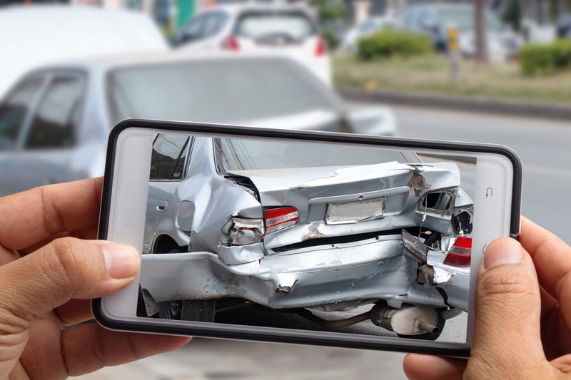 Hands hold a smartphone, photographing the severely damaged rear bumper and trunk of a silver car after an accident.