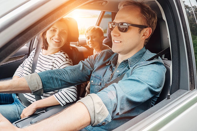 Family in a car, man driving in sunglasses, woman and child smiling in sunny car.