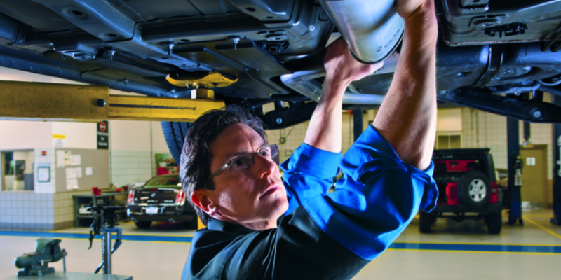 Mechanic in glasses and blue shirt working on the exhaust pipe of a car raised on a lift in a garage.