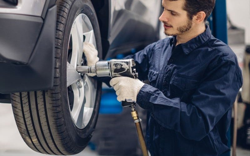 Mechanic in a blue shirt and gloves uses an impact wrench to work on a lifted car tire.